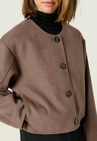 Brun, kort uldblandingsjakke med rund halsudskæring, der har fem sorte knapper og en glat tekstur. Sort turtleneck under.