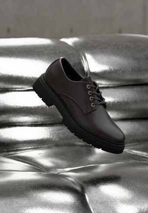 Mustast nahast paeltega king hõljub hõbedase läikiva pehmet nahkpinda imeva taustal, fookus on kinga külje ja talla detailidel.