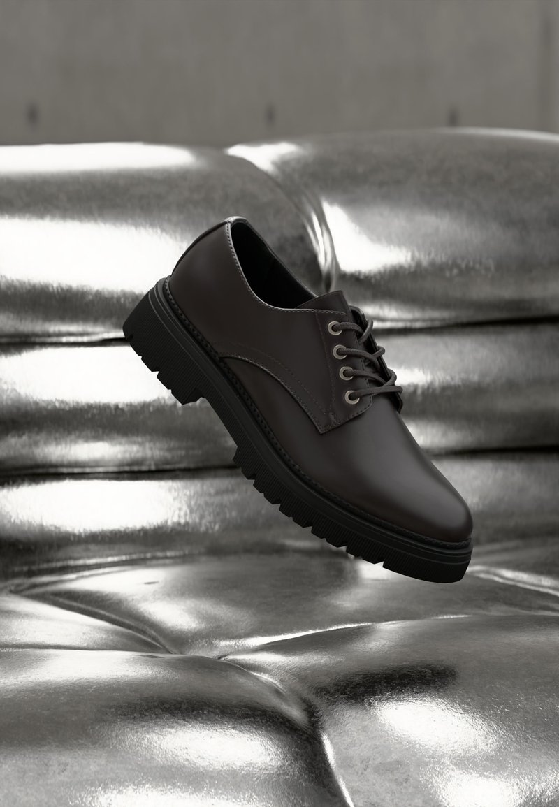 Μαύρο δερμάτινο παπούτσι δέσιμο με κορδόνια να αιωρείται πάνω από γυαλιστερή ασημί δερμάτινη επιφάνεια με καπιτονέ, εστιασμένο στις λεπτομέρειες της πλευράς και της σόλας του παπουτσιού.