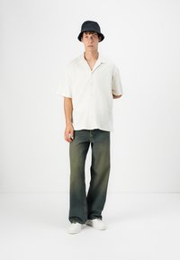 Kortærmet, struktureret hvid skjorte med knapper, parret med falmede grønne jeans og hvide sneakers. En sort bøttehat fuldender outfittet.
