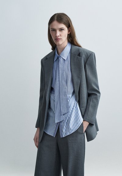 Jeune femme aux longs cheveux bruns portant un costume gris ample et une chemise rayée bleue et blanche superposée, debout devant un fond clair uni.