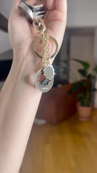 Furla sleutelhanger met een gouden ketting, een ronde hanger met bloemmotief uitsnijding en een ronde, gepolijste tag. Zilverkleurig beslag en een decoratieve ring.