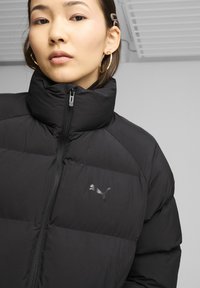 Sort pufferjakke med høj krave, lynlås foran og et diskret Puma-logo på brystet. Glat tekstur, isoleret design.