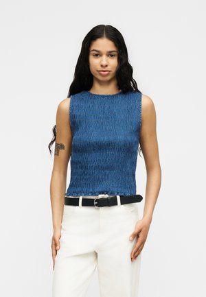 Fiatal nő áll, kék, ujjatlan, texturált felsőt visel, fehér nadrágot fekete övvel, és hosszú hullámos sötét hajat.