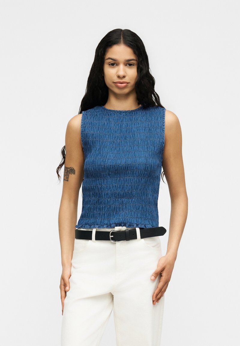 Jeune femme debout portant un haut bleu texturé sans manches, un pantalon blanc avec une ceinture noire, et de longs cheveux ondulés foncés.