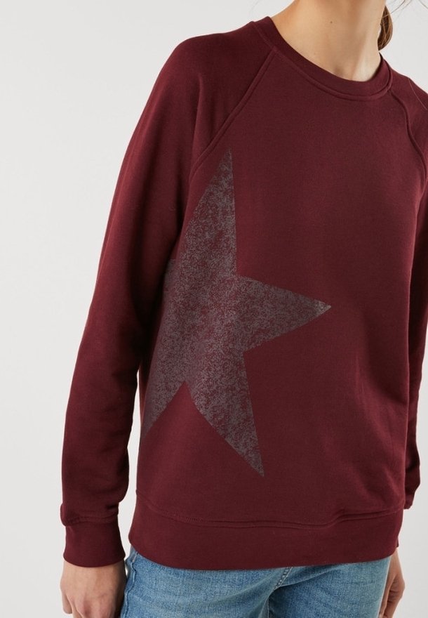Personne portant un sweat-shirt bordeaux à manches longues avec un grand motif d'étoile sombre sur le devant, associé à un jean bleu.