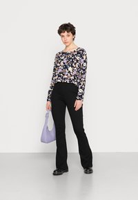 Langarmshirt mit Blumenmuster in Schwarz, Lila und Creme; kombiniert mit schwarzen Schlaghosen und einer helllila strukturierten Handtasche.