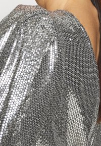 Silverfärgad paljettväv med en strukturerad, reflekterande yta. Har en rynkad halsringning och en lös passform som visar upp en modern design.