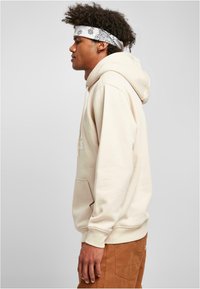 Beige kapucnis pulóver kenguruzsebből, húzózsinóros kapucnival és bordázott mandzsettával. Világosbarna nadrágon viselve, mintás fehér kendővel.