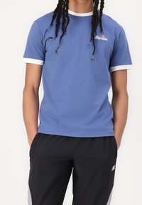 Modré bavlněné tričko s bílým žebrovaným límcem a rukávy, s malým logem na hrudi. Kombinováno s černými sportovními kalhotami.