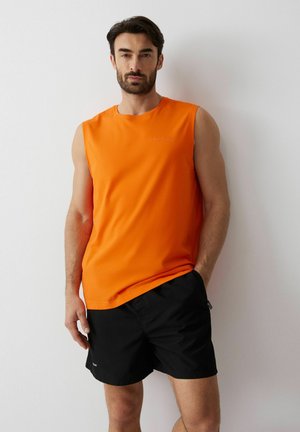 Mężczyzna w jaskrawo pomarańczowej koszulce bez rękawów i czarnych krótkich spodenkach stoi z jedną ręką w kieszeni na tle białej ściany.