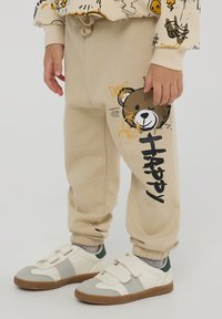 Beżowe dresy z bawełny z elastycznymi mankietami, z grafiką misia i napisem "Szczęśliwy" w czerni na lewym nogawce. Miękka faktura, luźny krój.