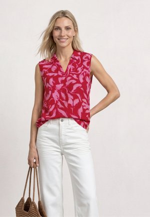 Femme souriante portant un chemisier sans manches rouge et rose avec un pantalon blanc, tenant un sac tressé marron dans une main, debout devant un fond uni.