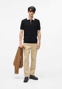 Crna polo majica s bijelim ovratnikom i rajsferšlusom, bež hlače, smeđi svečani sako od antilopa i crne cipele. Sunčane naočale upotpunjuju odijelo.