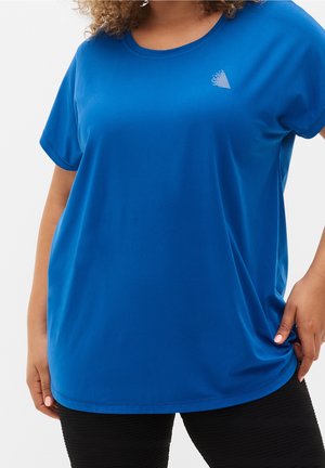 Lockig haarige Person, die ein locker sitzendes, hellblaues T-Shirt mit kurzen Ärmeln und kleinem weißem Logo auf der Brust sowie schwarze, strukturierte Hosen trägt.