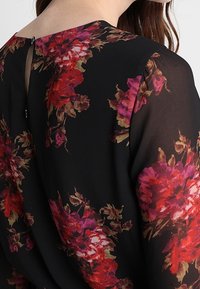 Blusa nera con motivo floreale in tonalità rosse e rosa, tessuto semi-trasparente e dettaglio a chiave sulla schiena. Texture morbida, vestibilità rilassata.