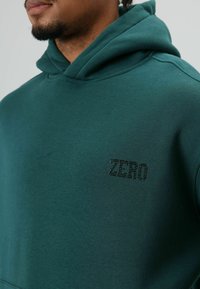 Sweat-shirt vert à capuche, avec un petit motif en texte noir "ZERO" en forme de clous sur le côté gauche de la poitrine. Texture lisse.