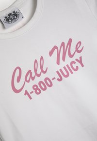 Balta medvilninė marškinėliai su rožiniais grafikos elementais, ant kurių parašyta "Call Me 1-800-JUICY". Marškiniai turi apvalią iškirptę ir prekės ženklo etiketę šalia apykaklės.