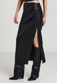 Femme portant une jupe midi en simili cuir noir avec des détails froncés sur le côté et une fente haute, associée à des bottes noires montantes à bout pointu.