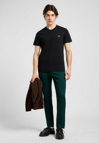 Schwarzes T-Shirt mit kurzen Ärmeln und kleinem weißen Logo, kombiniert mit grünen Hosen und einem braunen Strickpullover in der Hand. Schwarze Schuhe vervollständigen das Outfit.