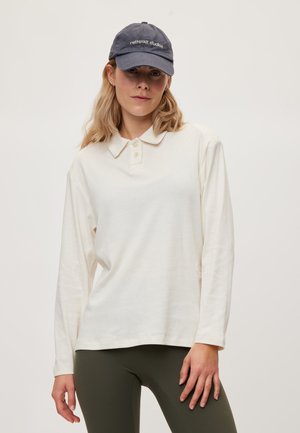 Kvinde iført en hvid langærmet poloshirt, olivengrønne bukser og en mørkeblå kasket med "rethinkit studios" tekst, står mod en ensfarvet baggrund.