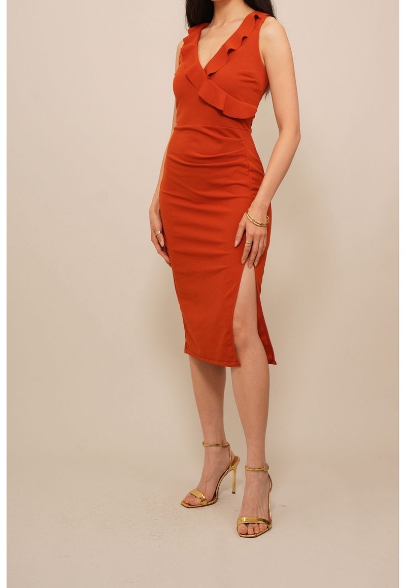 Femme portant une robe midi sans manches couleur rouille avec un col à volants et une fente haute à la cuisse, associée à des sandales dorées à talons hauts.