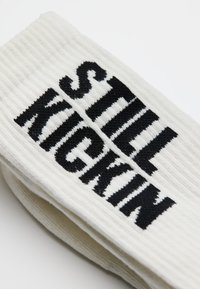 Witte geribde sokken met zwarte geborduurde tekst die "STILL KICKIN" zegt. De sok heeft een zachte textuur en een gemiddelde dikte.
