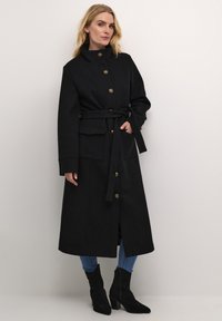 Manteau long noir avec des boutons dorés, taille ceinturée, poches latérales et col montant. Le mannequin porte un jean bleu et des bottines noires.