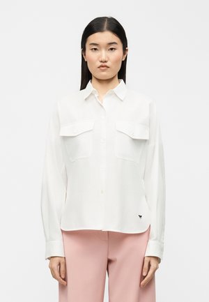 Jeune femme aux longs cheveux noirs portant une chemise blanche à boutons avec poches poitrine et un pantalon rose clair, debout devant un fond uni.