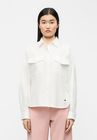 Jeune femme aux longs cheveux noirs portant une chemise blanche à boutons avec poches poitrine et un pantalon rose clair, debout devant un fond uni.