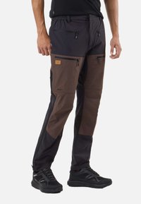 Man draagt zwarte en bruine outdoorbroek met meerdere ritszakken en zwarte sportschoenen, staand tegen een witte achtergrond.