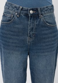 Pogled od blizu na modre jeanske hlače srednje pranja z gumbi spredaj, zankami za pas in vidnimi sprednjimi žepi, oblečene z modrim rebrastim topom.