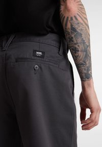 Pantalons en tissu gris foncé avec une poche arrière boutonnée et une étiquette logo Vans. Présente une texture lisse et un design à coupe classique.