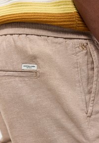 Primo piano della tasca di pantaloni beige con etichetta Scotch & Soda e logo ricamato, indossati con maglia gialla a maglia.