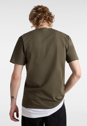 T-shirt olive avec des manches courtes, un col rond et une texture lisse, superposé sur une chemise blanche au niveau de l'ourlet.