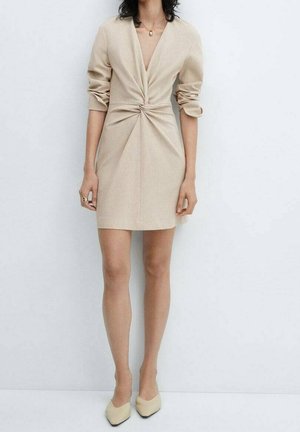 Femme portant une robe beige à col en V avec détail de taille froncée, manches retroussées, ballerines crème pointues et un collier pendentif en or.