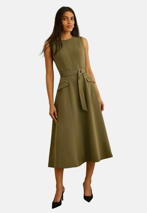 Kobieta w oliwkowej sukience midi bez rękawów z przednimi kieszeniami z klapami, paskiem w talii i czarnymi szpilkami z ostrym noskiem, stojąca na białym tle.