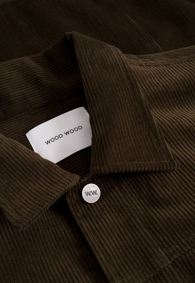 Cămașă din corduroy maro cu guler, având un etichetă albă pe care scrie „WOOD WOOD” și un buton metalic gravat cu „W.W.”.