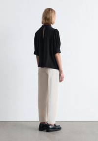 Schwarze, kurzärmelige Bluse mit hohem Kragen und Schlüssellochdetail auf der Rückseite, kombiniert mit lockeren beigen Hosen und schwarzen Schuhen.