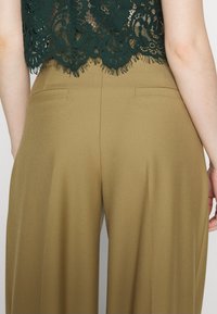 Hoge taille, wijde pijpen broek van khaki stof. Kenmerken zijn onder andere twee achterzakken en een gladde textuur. Gecombineerd met een groene kanten top.
