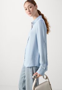 Camicia button-up azzurro chiaro, con texture, colletto e maniche lunghe, abbinata a jeans di denim chiaro. Tiene in mano una borsa color crema.