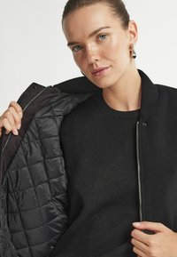 Svart bomberjacka med vadderad insida, ribbad krage och silverdragkedja. Den släta yttertyget och den texturerade foilen skapar kontrast.