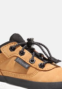 Světlé semišové tenisky s černými akcenty, texturovaným materiálem, elastickými tkaničkami a gumovou podrážkou s logem na boku.