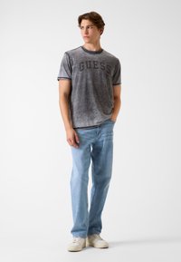 Szara koszulka z czarnym logo "GUESS", zestawiona z jasnoniebieskimi jeansami i białymi sneakersami, na tle o neutralnym kolorze.