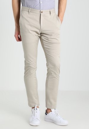 Man wearing beige slim-fit pants, light blue button-up shirt tucked in, and white sneakers with black detailing.