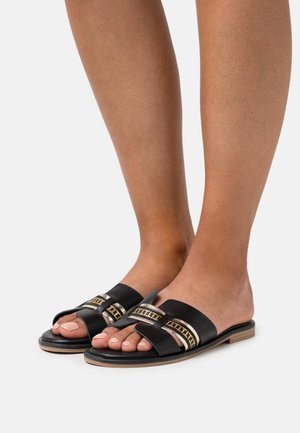 Musta nahkainen liukusandaali, jossa on kaksinkertaiset hihnat kullanvärisin koristuksin, litteä pohja ja avoin varvasosa.