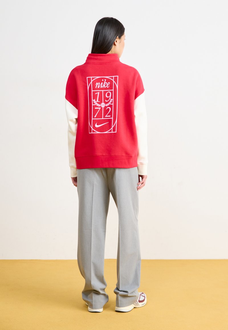 Červená mikina s bielou grafikou, ktorá obsahuje nápisy "nike," "1972" a logo. Dlhé svetlošedé nohavice dopĺňajú outfit. Mäkka, pohodlná tkanina.