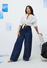 Jean en denim sombre à jambes larges associé à une chemise blanche nouée. Texture lisse, coutures contrastantes et coupe décontractée.