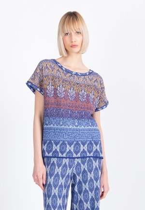 Donna con taglio bob biondo che indossa una maglietta a maniche corte con fantasia multicolore e pantaloni abbinati con motivo blu, in piedi su sfondo bianco.