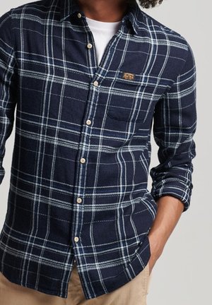 Man wearing a navy blue and white plaid button-up shirt with rolled sleeves and beige pants, hand in pocket.